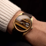 Vintage Rolex Datejust 1601 Tiger Eye de 1974 sur un poignet. 36mm avec boîtier en or jaune 18K, cadran en pierre Tiger Eye, mouvement automatique et bracelet en cuir.