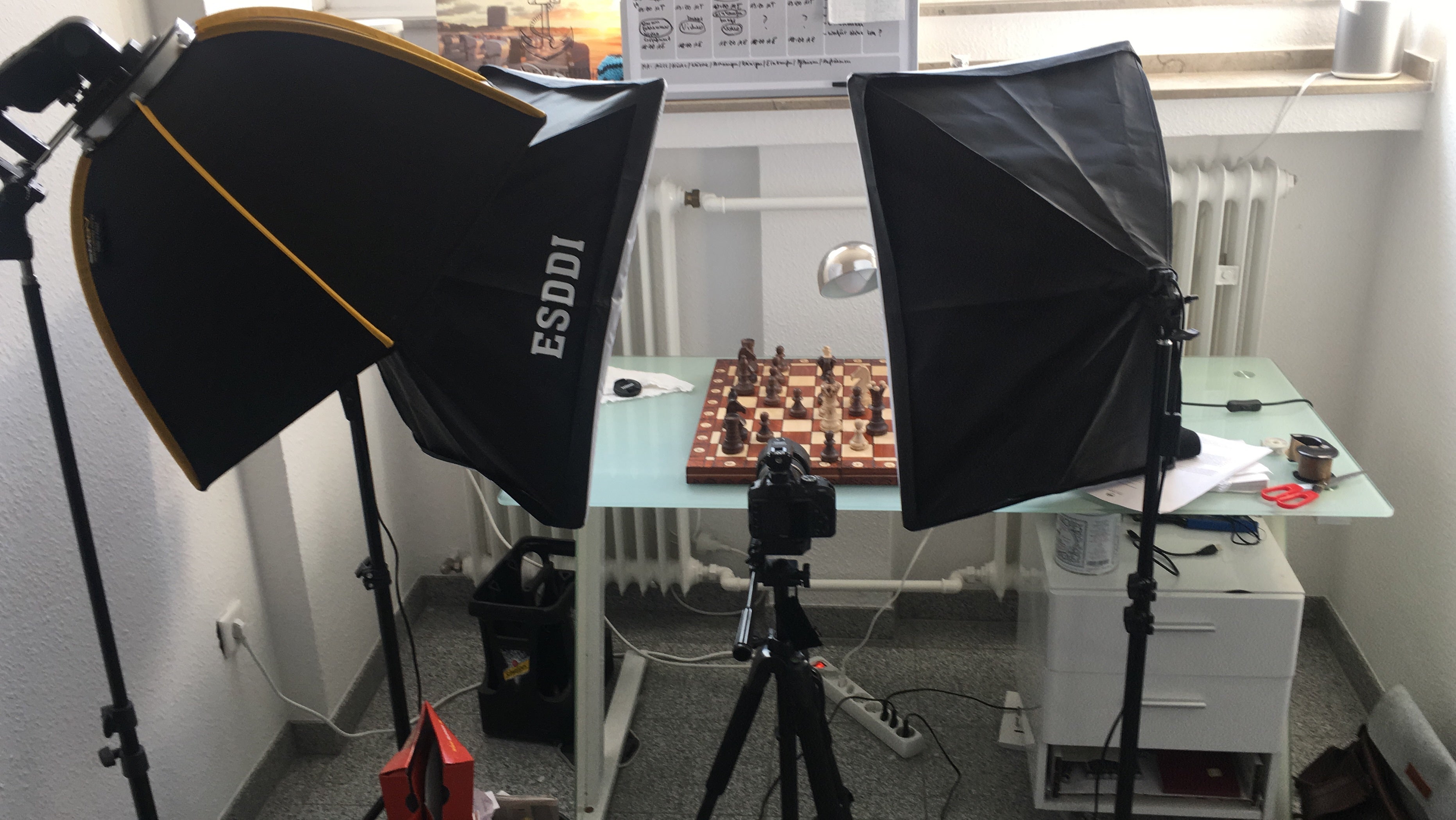 Setup fotografico con due luci softbox, una fotocamera su un treppiede e una scacchiera su una scrivania di vetro.