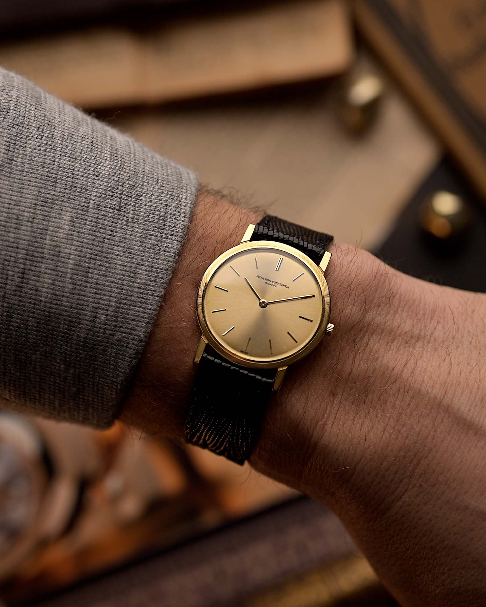 ساعة Vacheron Constantin Patrimony 6351 عتيقة الطراز بعلبة مستديرة رفيعة من الذهب عيار 18 قيراط، وميناء ذهبي، وتشغيل يدوي، وحزام أسود على المعصم على خلفية ضبابية