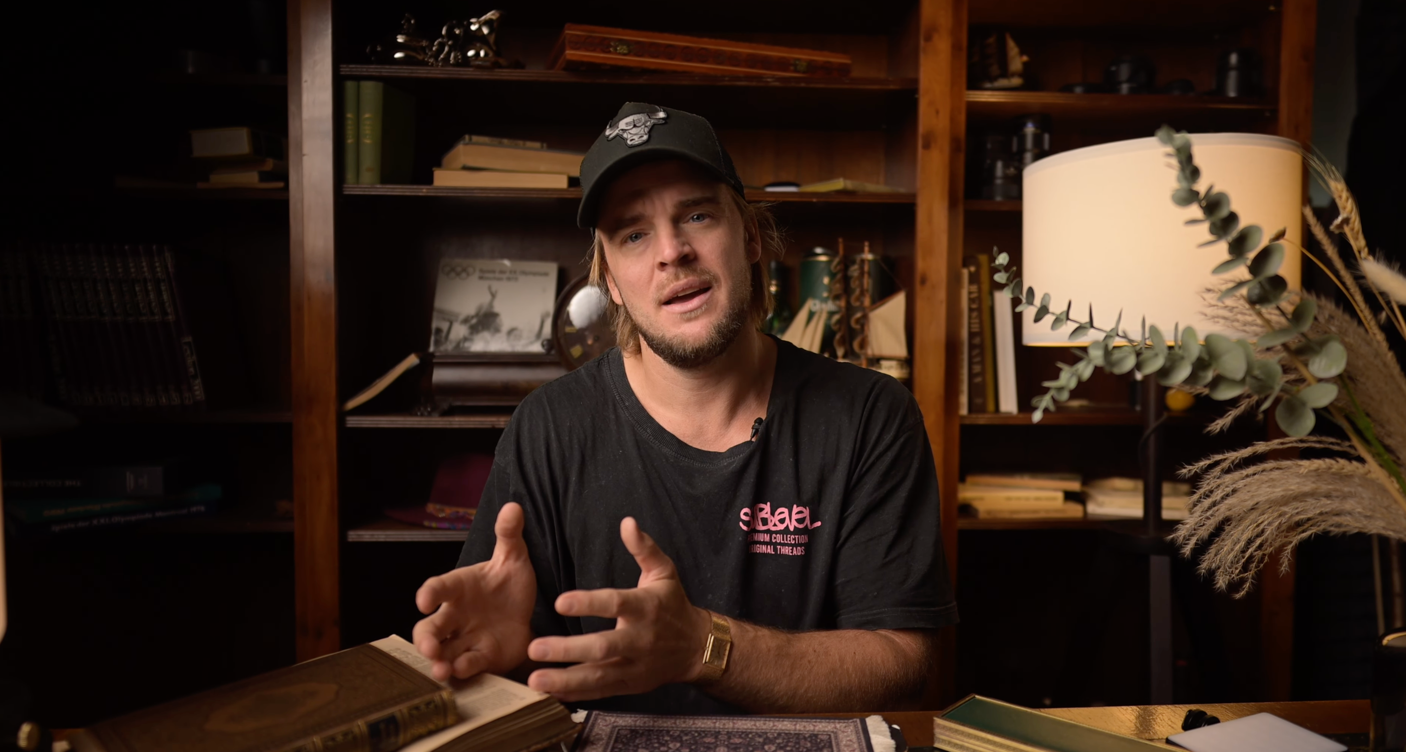 Man in a dark t-shirt and cap sitting at a desk with a bookshelf in the background