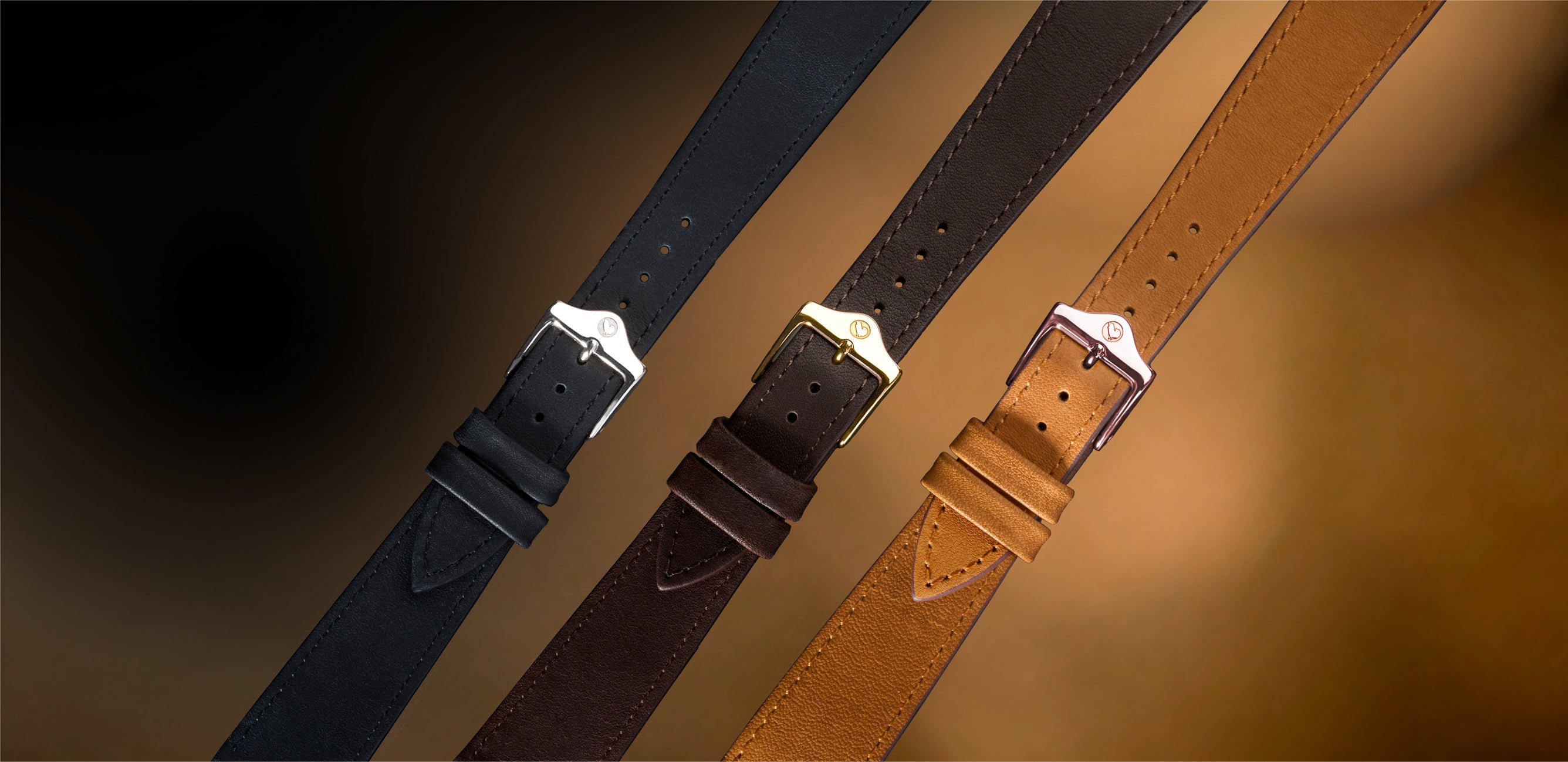 Three leather watch straps in black, dark brown, and tan, displayed diagonally on a soft, warm gradient background.