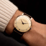 Rosegold Vintage Jaeger LeCoultre 18k Rose Railroad watch from the 1950s on a person's wrist with a dark background. 35mm wide with 18k Rose Gold Case, Manual Wind Caliber P450/4C, Cream Dial and Brown Leather Strap.