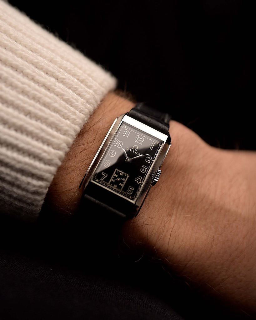 Vintage Omega Rectangular Syringe watch, black gilt dial, stainless steel case, manual movement, on black leather strap, on a wrist on black background