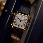 Vintage Cartier Ceinture 78099 Uhr mit quadratischem Gehäuse aus 18 Karat Gelbgold, weißem Zifferblatt, Handaufzug und blauem Lederarmband auf unscharfem Hintergrund.