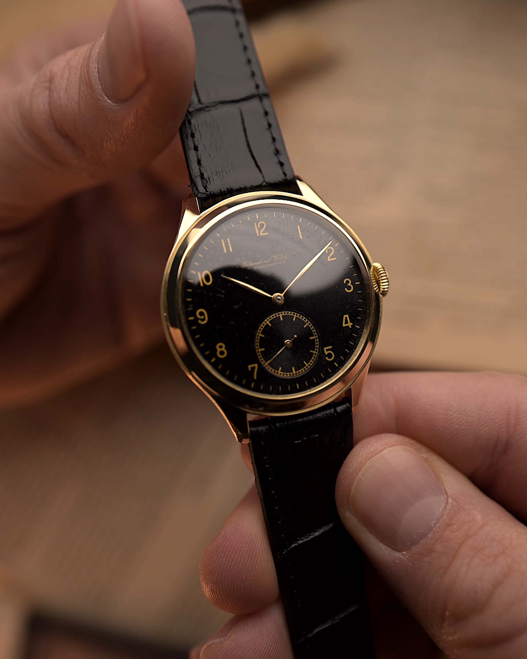 Vintage IWC Pre Hermet Uhr, aus 14 Karat Gelbgold, schwarz vergoldetes Zifferblatt, Cal 83 Handaufzug, schwarzes Lederarmband, in den Händen vor unscharfem Hintergrund gehalten.