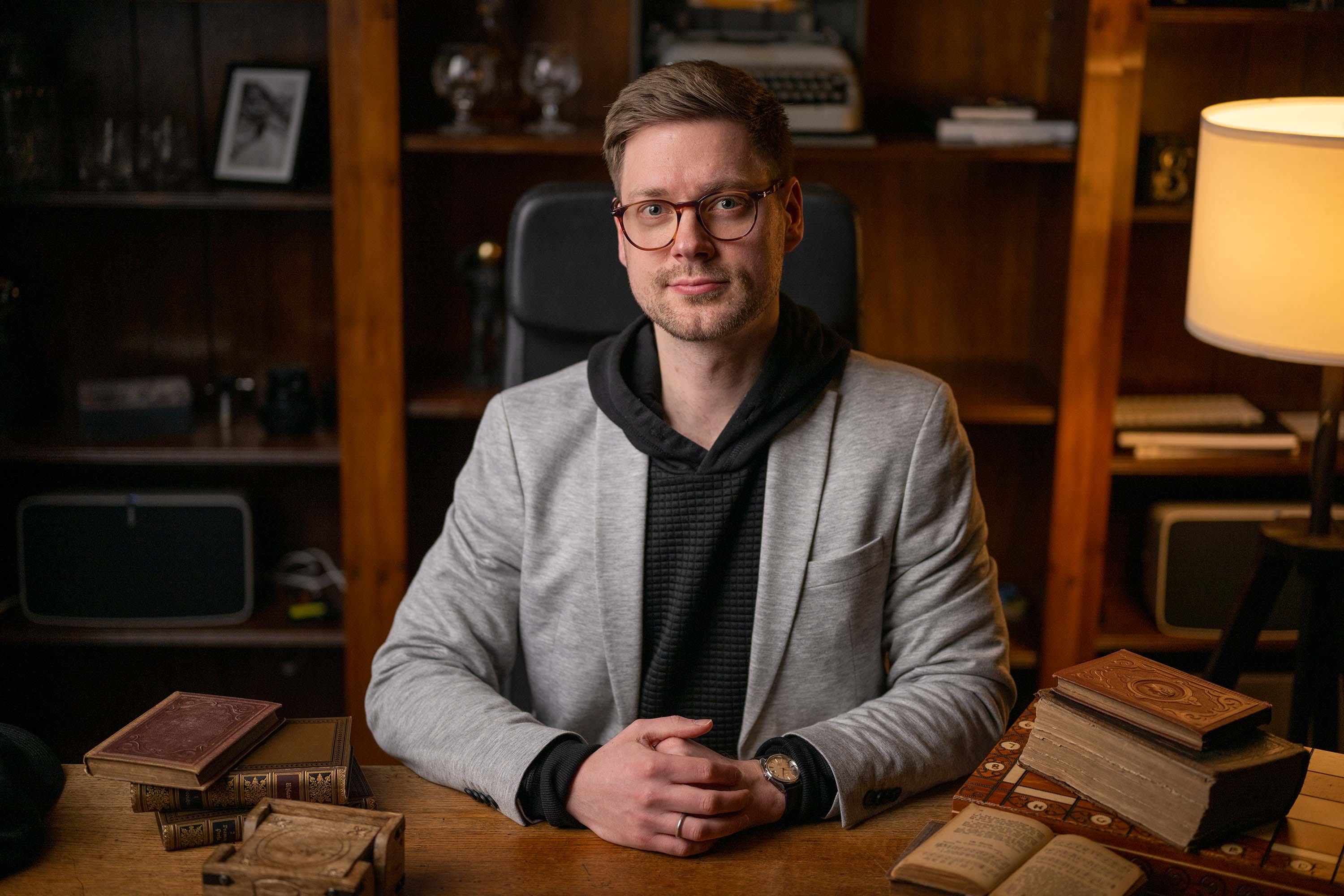 Ein Mann mit Brille, grauem Blazer und schwarzem Kapuzenpulli sitzt an einem Tisch.