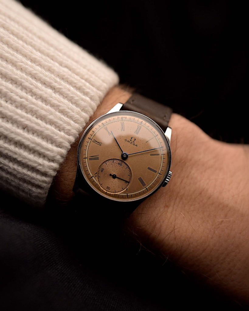 Vintage Omega Art Deco Uhr mit lachsfarbenem Zifferblatt, Edelstahlgehäuse, Handaufzugswerk und braunem Lederband am Handgelenk auf schwarzem Hintergrund