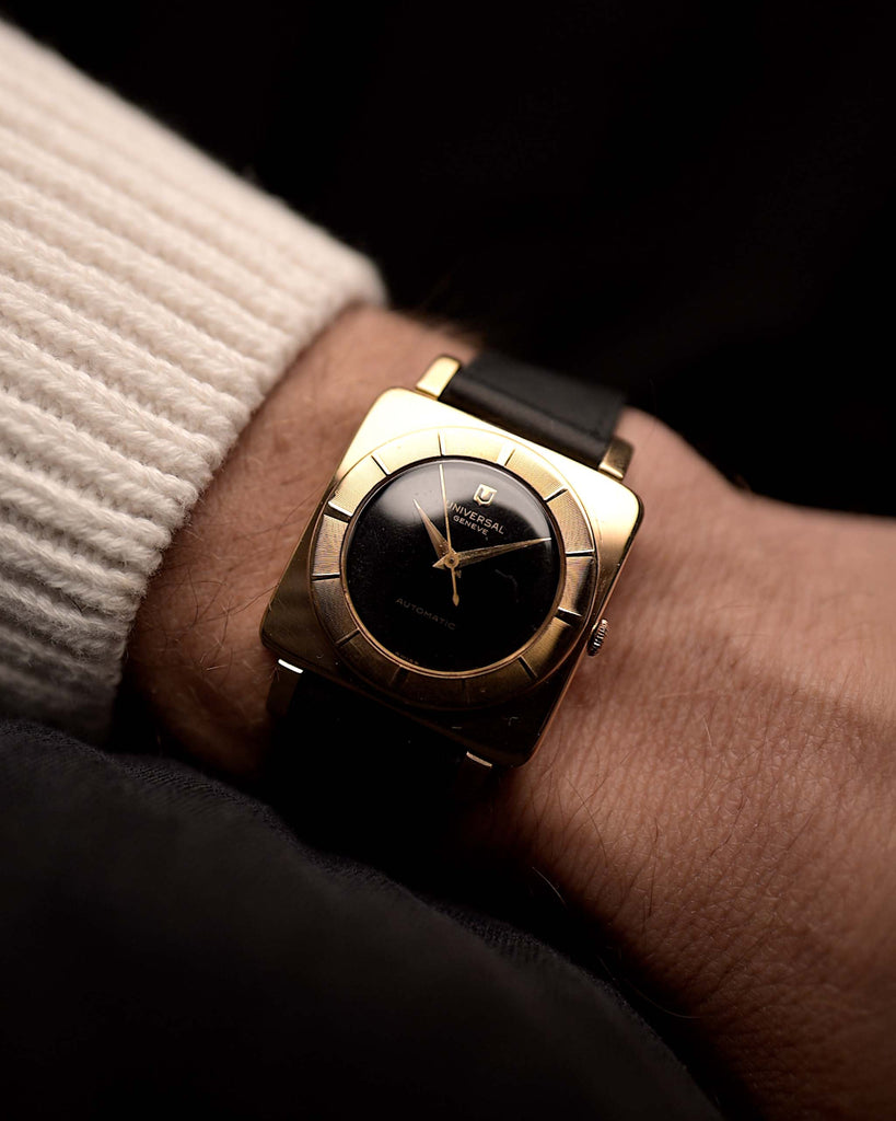 Universal Genéve 10237 Cioccolatone von 1956 an einem Handgelenk auf schwarzem Hintergrund. 30 mm breit mit Gehäuse aus 18 Karat Roségold, Uhrwerk mit Handaufzug, schwarzem Zifferblatt und schwarzem Lederarmband.