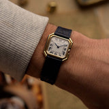 Reloj vintage Cartier Ceinture 78099 con caja cuadrada de oro amarillo de 18 quilates, esfera blanca, cuerda manual y correa de cuero azul sobre fondo difuminado.