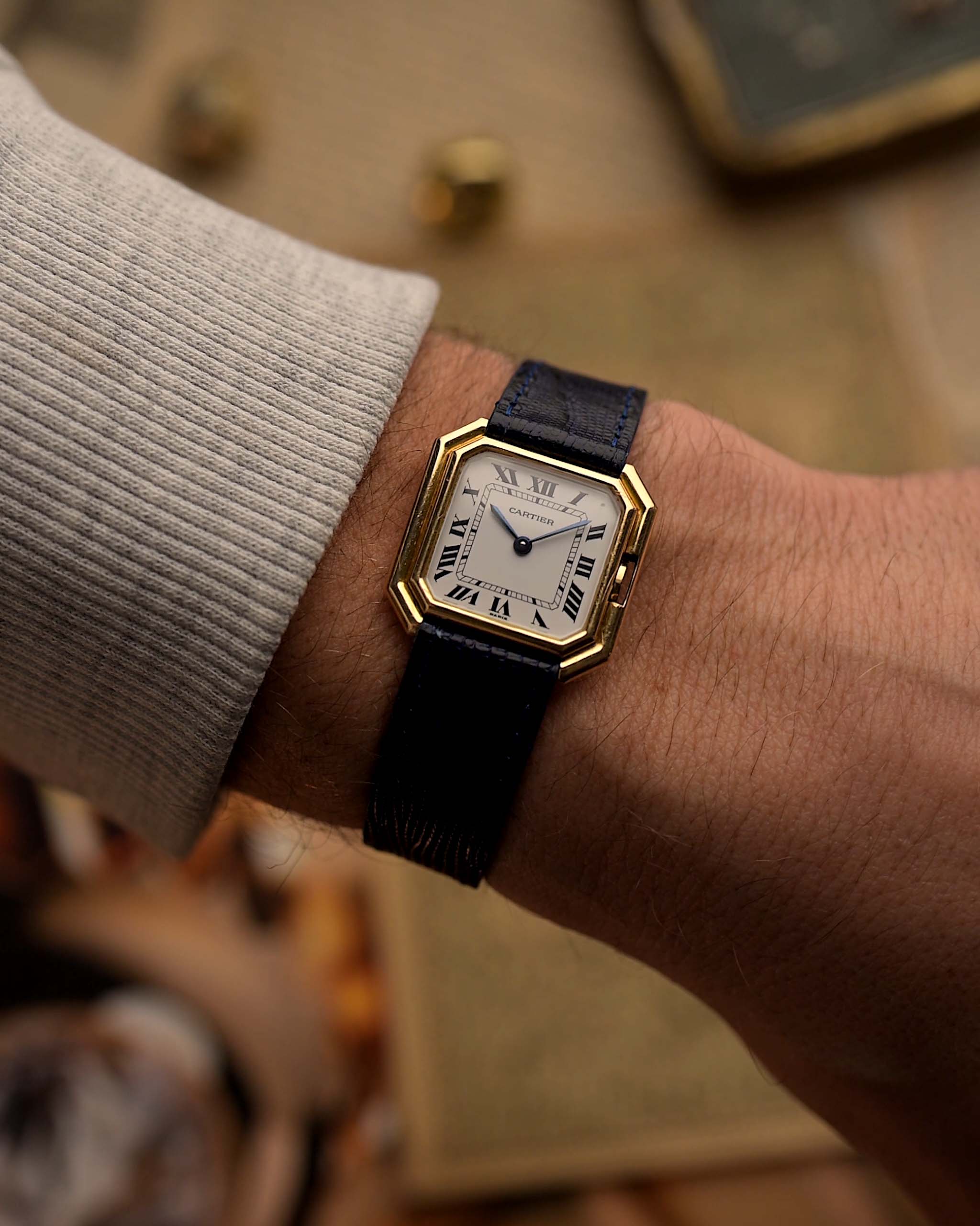 Reloj vintage Cartier Ceinture 78099 con caja cuadrada de oro amarillo de 18 quilates, esfera blanca, cuerda manual y correa de cuero azul sobre fondo difuminado.