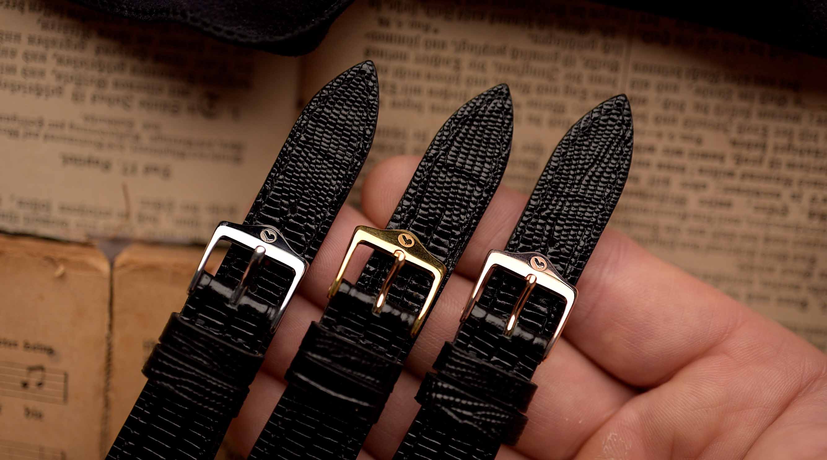Tres correas de reloj negras con diferentes hebillas sujetas por una mano sobre una superficie texturizada.