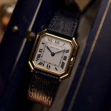 Montre vintage Cartier Ceinture 78099 avec boîtier carré en or jaune 18 carats, cadran blanc, remontage manuel, bracelet en cuir bleu sur fond flou.