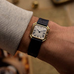 Montre vintage Cartier Ceinture 78099 avec boîtier carré en or jaune 18 carats, cadran blanc, remontage manuel, bracelet en cuir bleu sur fond flou.