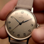 Montre Omega 2325 Jumbo Leaf vintage, cadran argenté, boîtier en acier inoxydable, bracelet en cuir gris, tenue à la main sur fond flou