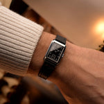 Montre Omega Rectangular Syringe vintage, cadran noir doré, boîtier en acier inoxydable, mouvement manuel, sur bracelet en cuir noir, au poignet sur fond flou