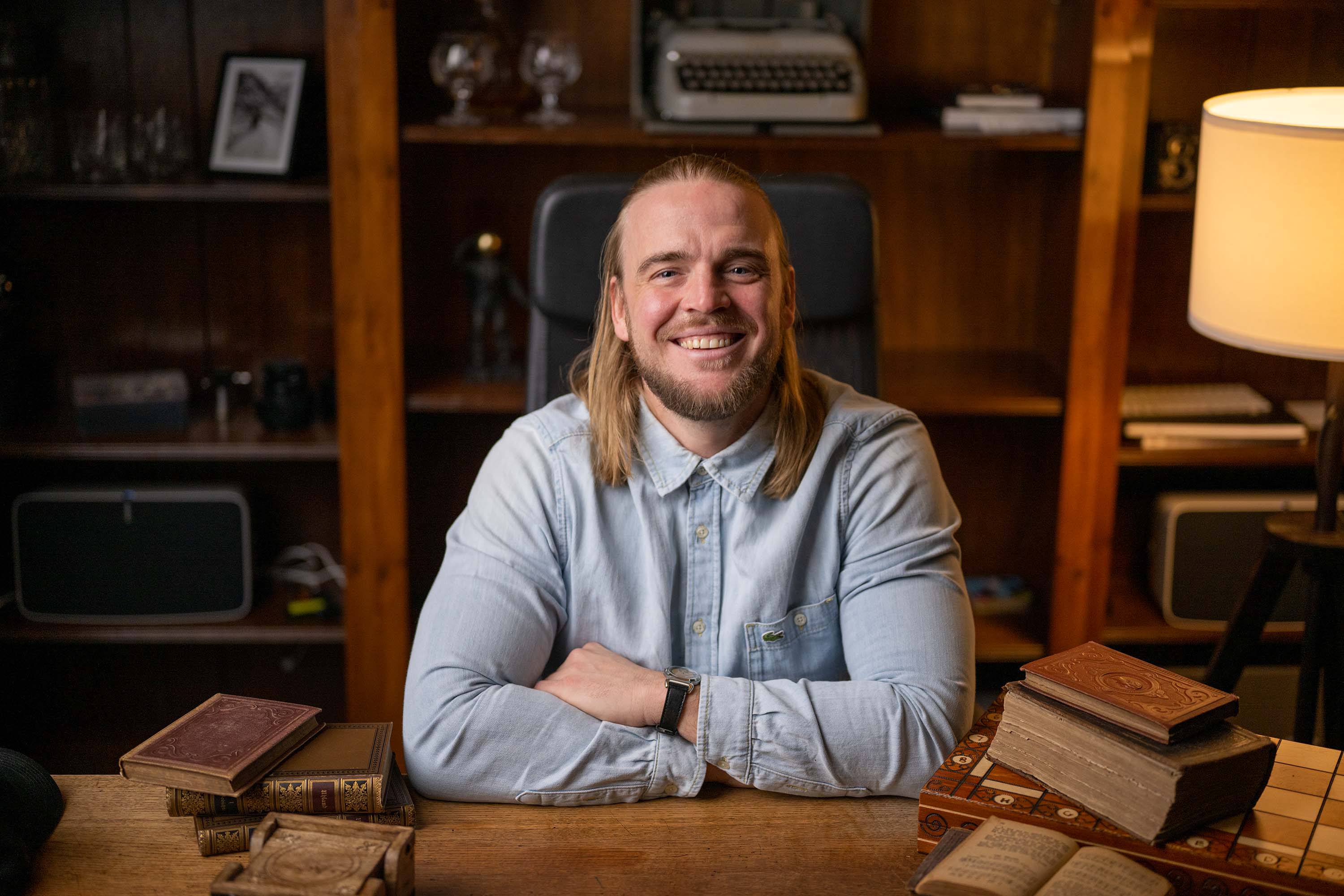 Homme souriant aux cheveux longs et à la chemise en jean, assis à une table.
