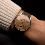 Montre vintage Omega Art Deco avec cadran saumon, boîtier en acier inoxydable, mouvement à remontage manuel et bracelet en cuir marron au poignet sur fond noir.