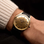 金色 Omega Constellation 168005 Date 復古腕錶戴在深色背景的人的手腕上。34mm 闊，18K 黃金錶殼，561 自動機芯，無瑕金色錶盤和黑色皮革錶帶。