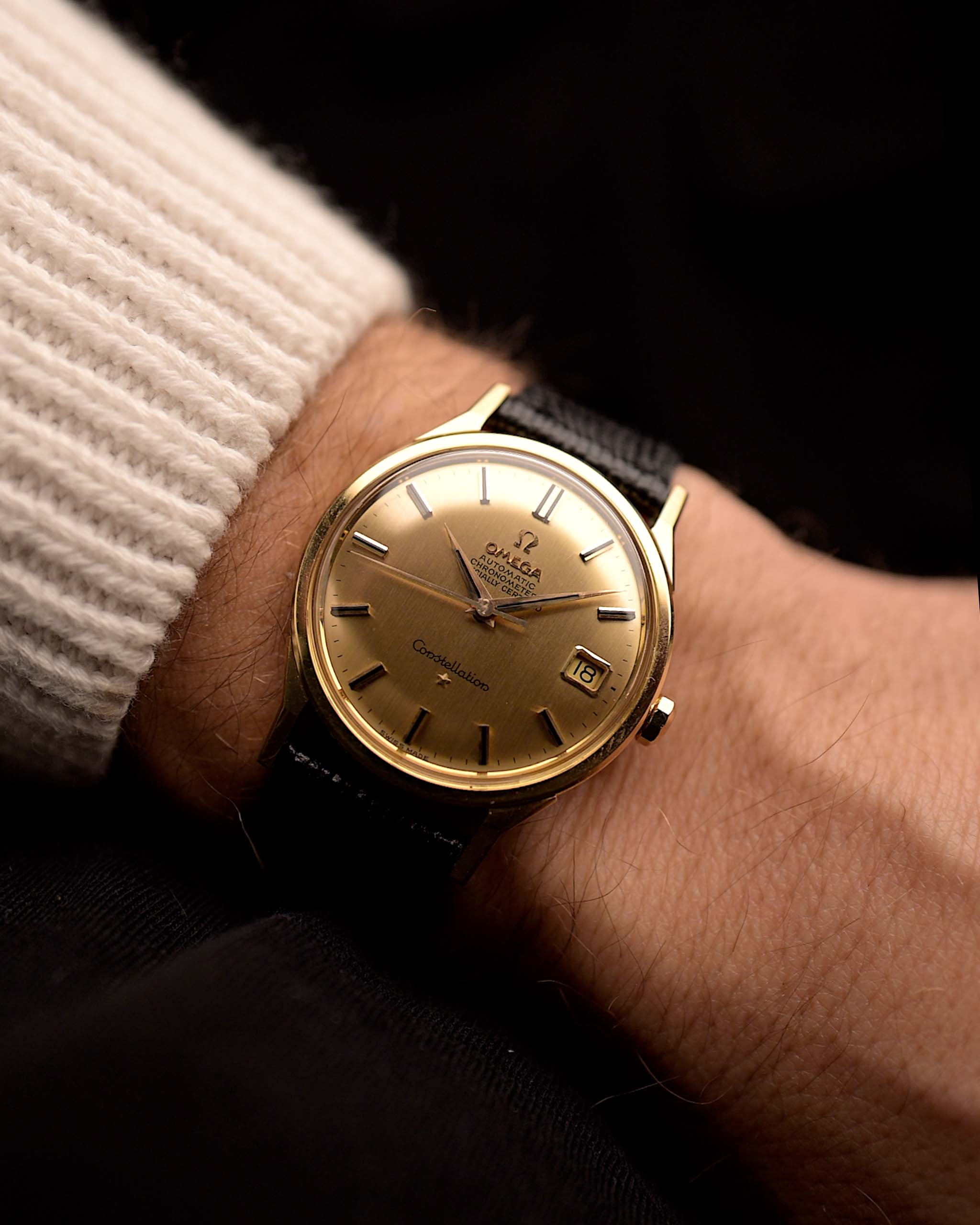 金色 Omega Constellation 168005 Date 復古腕錶戴在深色背景的人的手腕上。34mm 闊，18K 黃金錶殼，561 自動機芯，無瑕金色錶盤和黑色皮革錶帶。